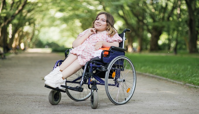 Portrait of pleasant young woman with spinal muscular atrophy sitting in a wheelchair and smiling on camera among green summer park